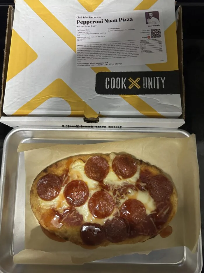 CookUnity pepperoni naan pizza with melted cheese on parchment on a metal baking tray, chef label on the open box behind