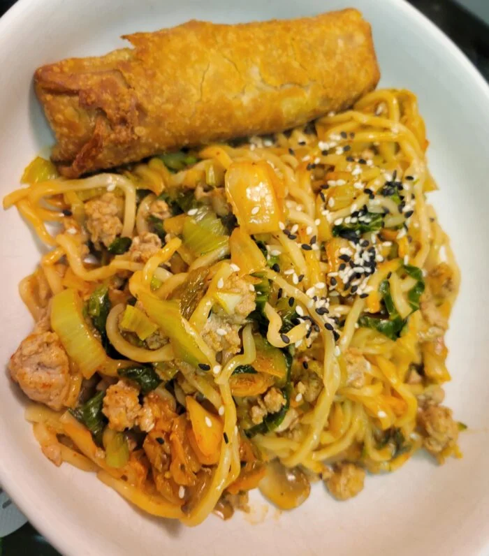 Blue Apron kimchi lo mein with ground pork, bok choy and sesame seeds, served with an egg roll in a white bowl