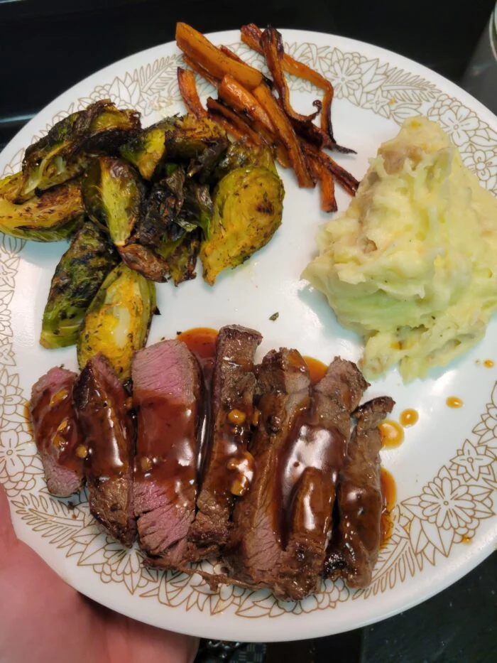Blue Apron sliced steak with steak sauce, cheesy mashed potatoes, roasted Brussels sprouts and carrots on a plate