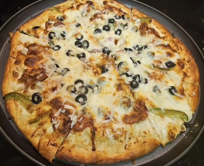 Blue Apron three cheese pizza with black olives, green peppers and grated cheese, sliced on a dark pizza pan