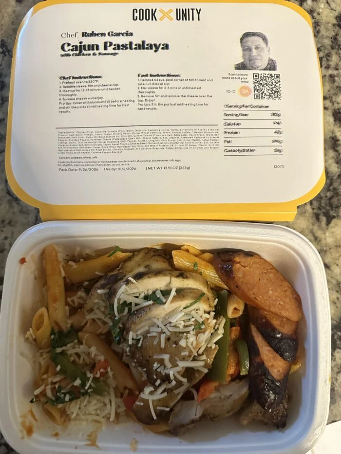 CookUnity Cajun pastalaya, penne with sliced chicken, smoked sausage, peppers and grated cheese in the tray, chef label open behind it