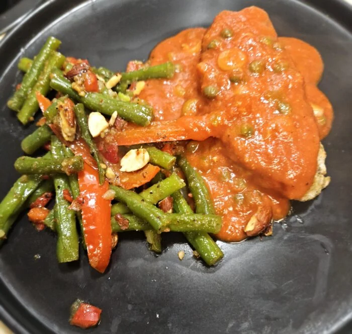 Green Chef pork chop under tomato sauce with peas, beside green beans, red pepper and sliced almonds on a black plate