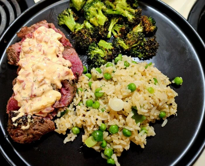 Green Chef sliced ranch steak with creamy red pepper sauce, rice with peas and roasted broccoli on a black plate