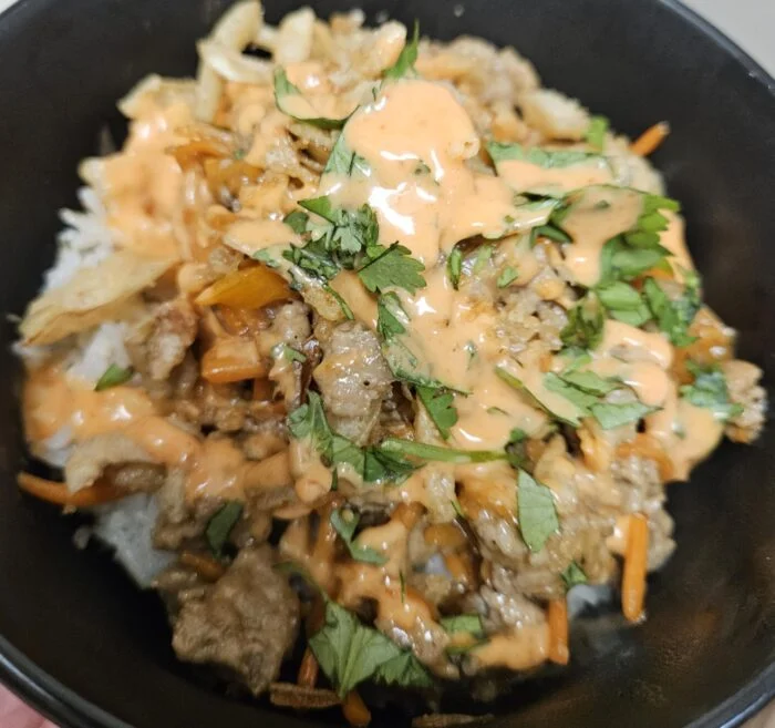 HelloFresh sesame pork bowl with sriracha mayo, cilantro, crispy onions and carrots over white rice in a black bowl