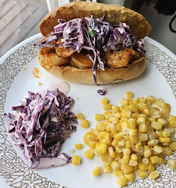 HelloFresh buttered shrimp roll topped with purple cabbage slaw, with extra slaw and corn on a patterned plate