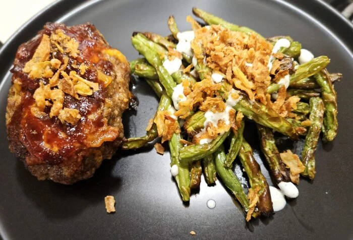 Home Chef bacon cheddar beef meatloaf with BBQ sauce and crispy onions, ranch green beans on a dark plate