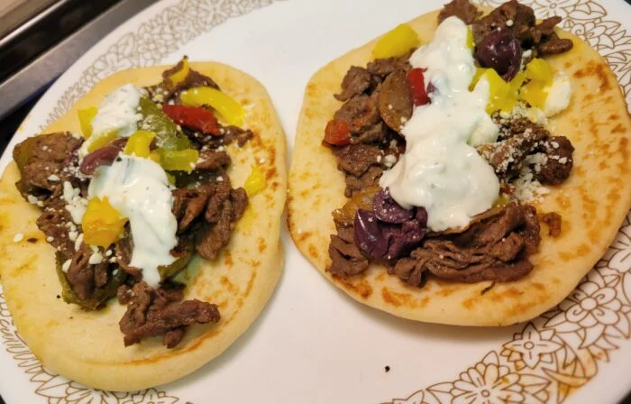 Home Chef Italian style beef gyros on two flatbreads with tzatziki, feta, olives and yellow and red peppers on a floral plate