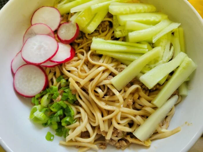 Marley Spoon Beijing style pork noodles with ground pork, cucumber sticks, sliced radish and scallions in a white bowl