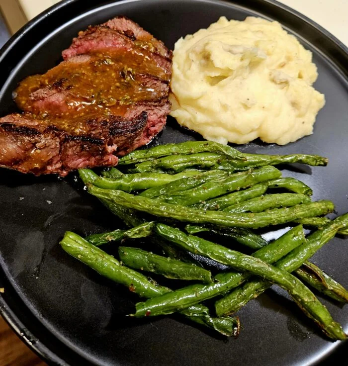 Marley Spoon honey Dijon glazed steak, sliced, with mashed potatoes and roasted green beans on a black plate
