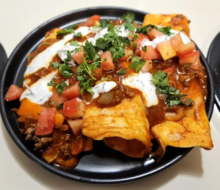Marley Spoon chorizo chili beef enchiladas topped with diced tomato, cilantro and crema drizzle on a black plate