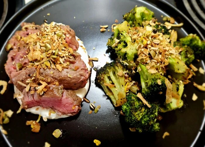 Marley Spoon sirloin steak, sliced over blue cheese crema with pepita gremolata, roasted broccoli on a black plate
