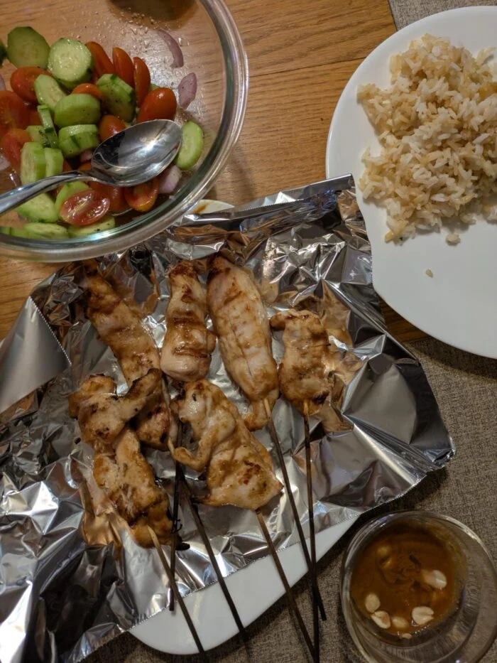 HelloFresh Thai chicken sate skewers resting on foil beside a bowl of rice and a cucumber salad