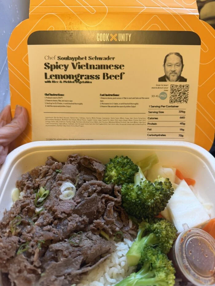 CookUnity spicy Vietnamese lemongrass beef over white rice with broccoli, pickled vegetables and a sauce cup, meal label held up behind the tray