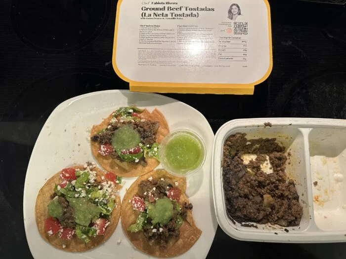 Three CookUnity ground beef tostadas plated with guacamole, tomato and a green salsa cup, beside the tray and chef label