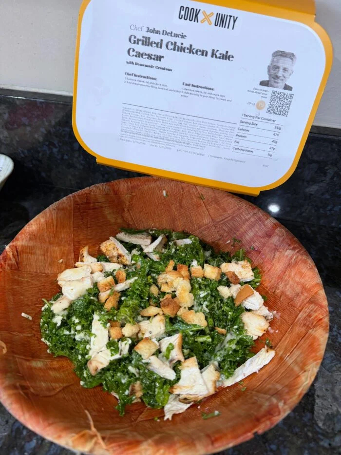 CookUnity grilled chicken kale Caesar salad with croutons in a wooden bowl, chef label behind