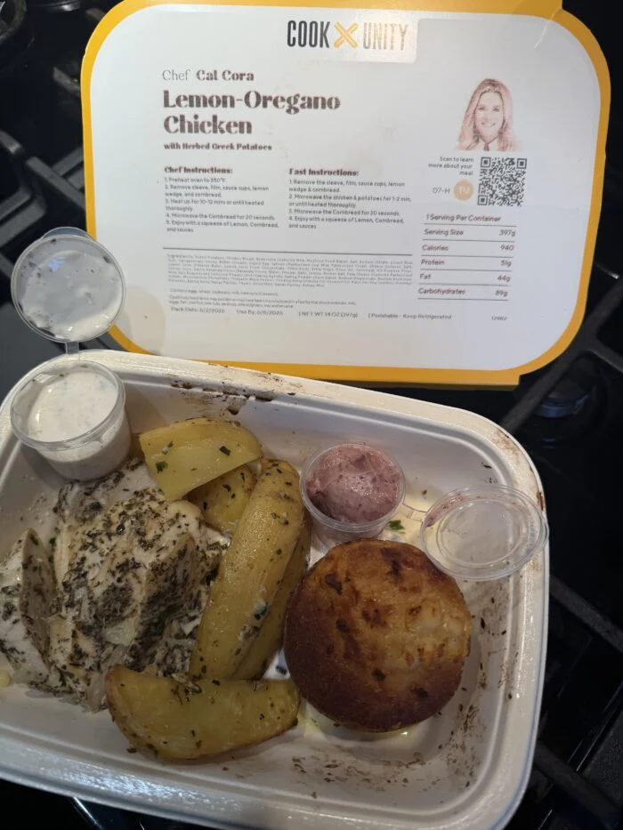 CookUnity lemon oregano chicken with roasted potatoes, a muffin and sauce cups in the tray, chef label behind